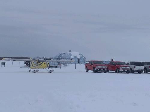 A small yellow aircraft parked on snowy ground, with red and white trucks nearby and a dome structure in the background.
