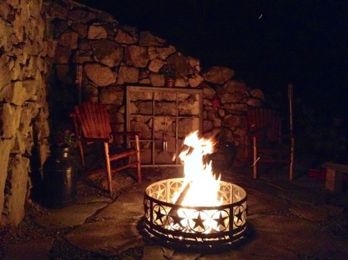 A cozy outdoor fire pit surrounded by stone walls and wooden chairs, illuminated at night.
