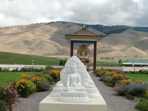 A serene landscape featuring white Buddha statues, colorful flowers, and a temple against a backdrop of rolling hills.