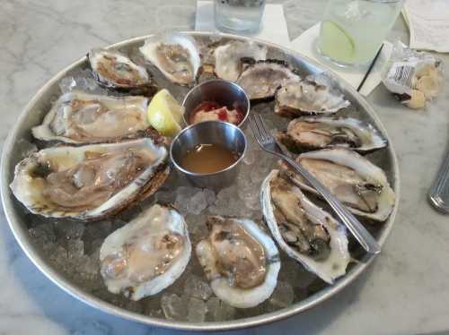 A platter of fresh oysters on ice, served with lemon, cocktail sauce, and a dipping sauce.