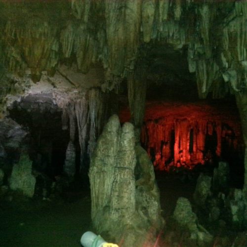 Stalactites and stalagmites in a dimly lit cave, with red lighting illuminating the rock formations.