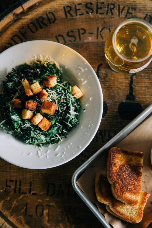 A bowl of kale salad topped with croutons and cheese, alongside a tray of toasted bread and a glass of drink.