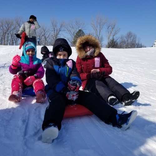 Children in winter clothing sit on sleds, ready to go down a snowy hill on a sunny day.