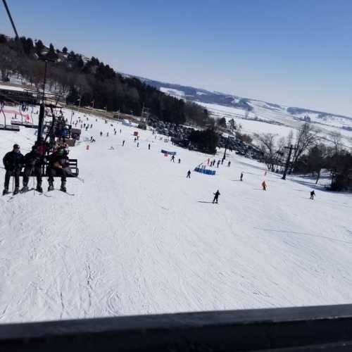 A snowy ski slope with skiers and snowboarders, viewed from a ski lift. Trees and hills in the background.