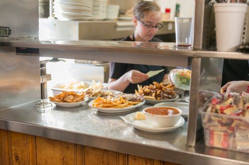 A busy diner kitchen with plates of food, including fries and soup, as a server prepares orders behind the counter.