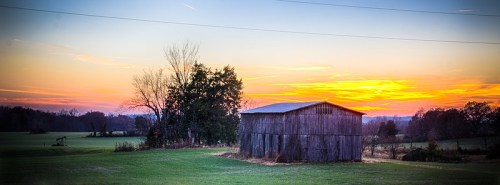 A rustic barn stands in a field at sunset, with vibrant orange and purple skies and trees in the background.