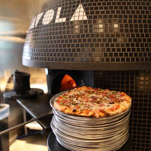 A stack of pizzas sits on a counter near a traditional pizza oven with a tiled exterior.