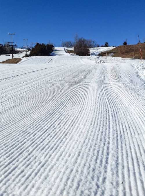 A snow-covered ski slope with freshly groomed tracks under a clear blue sky.