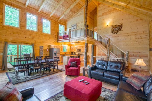 Cozy wooden cabin interior with a living area, dining table, kitchen, and staircase, surrounded by large windows.