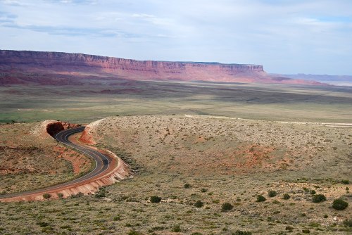 Winding road through a vast, arid landscape with red rock formations and distant cliffs under a cloudy sky.