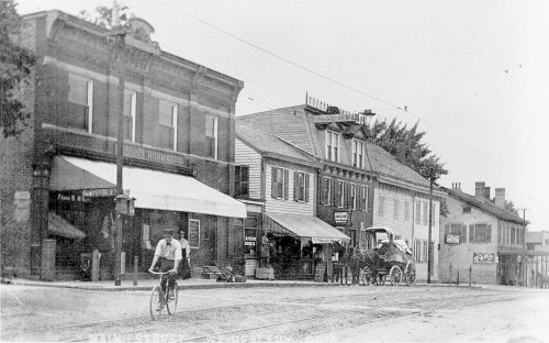 A black-and-white photo of a street scene featuring a cyclist, horse-drawn carriage, and historic buildings.