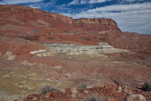 A rugged landscape featuring red rock formations, layered cliffs, and sparse vegetation under a partly cloudy sky.