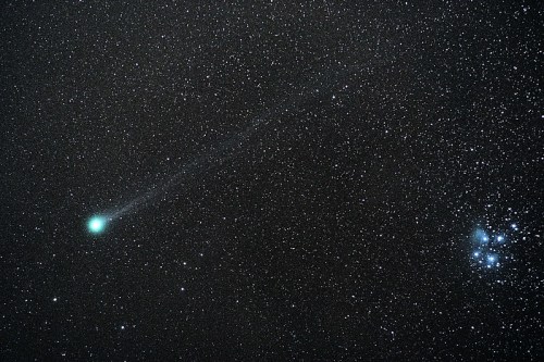 A bright green comet streaks across a starry night sky, near a cluster of stars on the right.