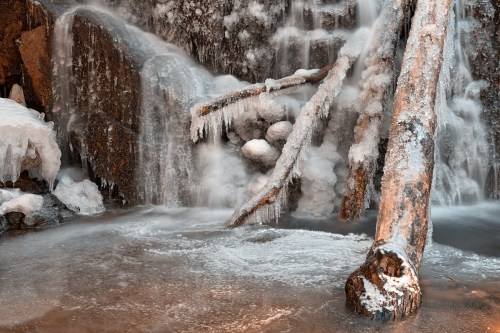A frozen waterfall with icicles hanging from rocks, surrounded by ice-covered logs and a misty, icy pool below.