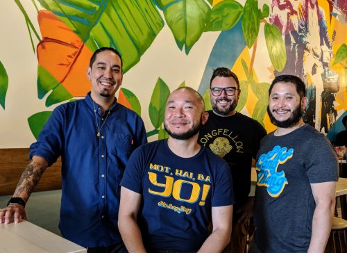 Four men stand together in a colorful restaurant, smiling and wearing casual shirts. Tropical plants decorate the background.