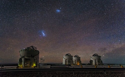 Three large telescopes under a starry sky, with the Milky Way and two bright clouds of Magellan visible above.