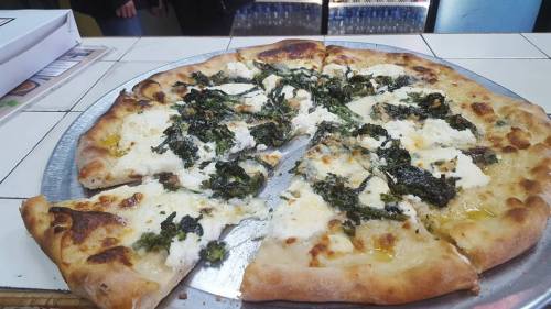 A pizza with a crispy crust topped with ricotta cheese and leafy greens, partially sliced on a metal tray.