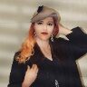 A woman with long, wavy hair wearing a stylish hat and earrings, posing confidently against a soft background.