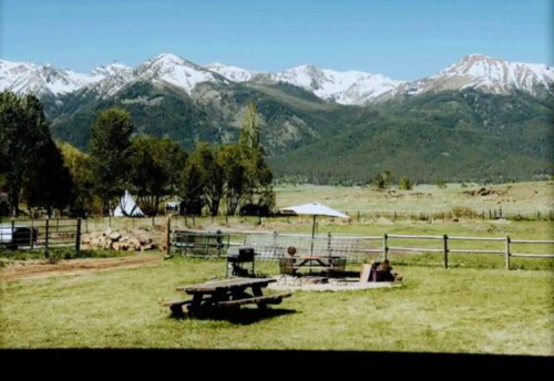 A scenic view of a grassy field with a picnic table, surrounded by mountains and trees under a clear blue sky.