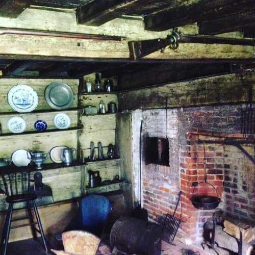 A rustic kitchen interior with wooden beams, shelves of dishes, and a brick fireplace.