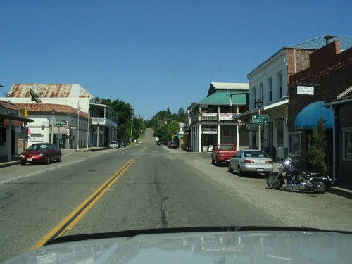 A quiet street scene with shops and parked cars under a clear blue sky. A motorcycle is visible on the right side.