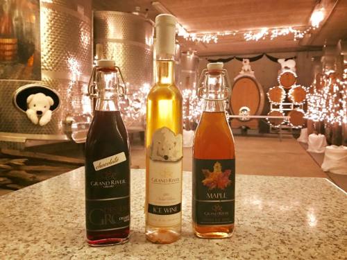 Three bottles of wine on a countertop, with a winery backdrop featuring barrels and festive lights.