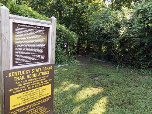 Sign at the entrance of Woods Walk Trail in Kentucky State Parks, surrounded by lush greenery and trail regulations.