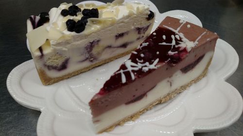 Two slices of layered cheesecake on a decorative plate, one topped with berries and white chocolate, the other with a berry glaze.
