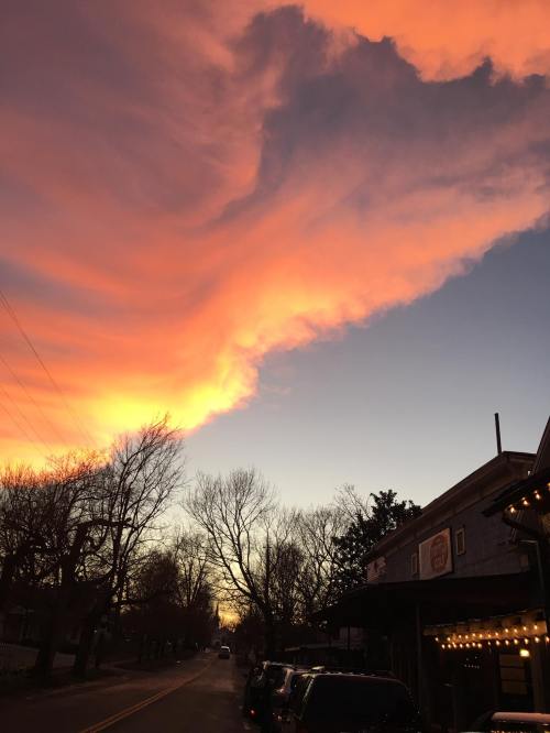 A vibrant sunset with orange and pink clouds over a quiet street lined with bare trees and buildings.