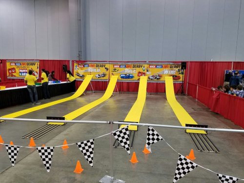 A colorful event setup with yellow ramps for a derby, featuring checkered flags and orange cones in a convention center.