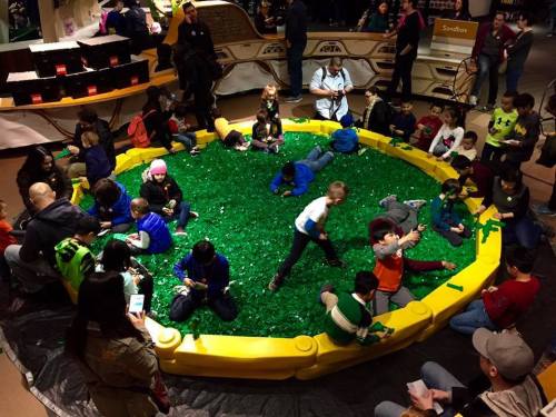 A group of children playing in a large, circular sandbox filled with green building blocks, surrounded by adults.
