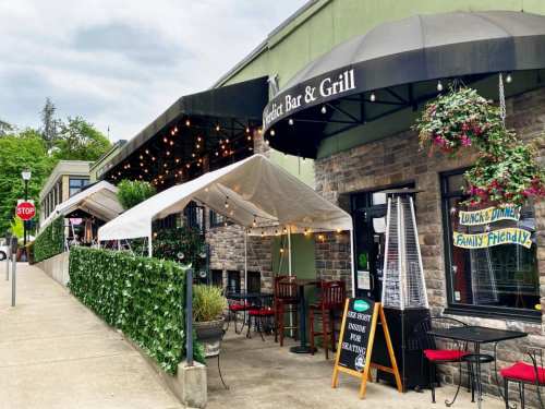Outdoor seating area of a bar and grill, featuring tents, string lights, and greenery along the sidewalk.