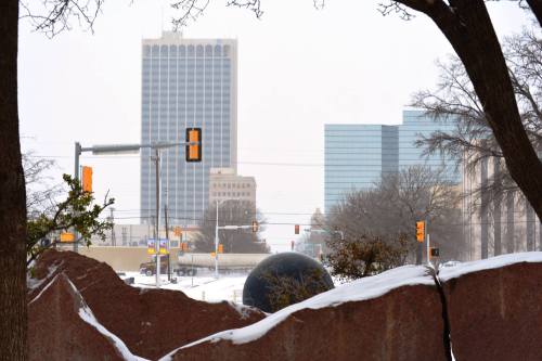 Snow-covered cityscape featuring tall buildings and a large stone sculpture in the foreground.