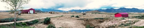 Panoramic view of a rural landscape with red barns, mountains in the background, and a dirt road.