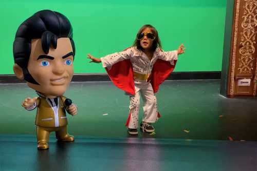 A toy figure of Elvis stands beside a child in a white jumpsuit and sunglasses, posing dramatically on a stage.