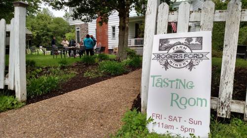 A sign for the Rogers Ford Tasting Room outside a farm, with people gathered in the background near a house.