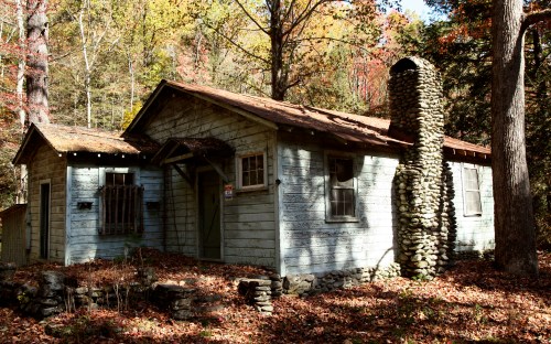 A rustic, weathered cabin surrounded by autumn foliage and a stone chimney, nestled in a wooded area.