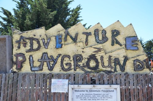 A colorful wooden sign reading "Adventure Playground" at the entrance of a playground, surrounded by trees.