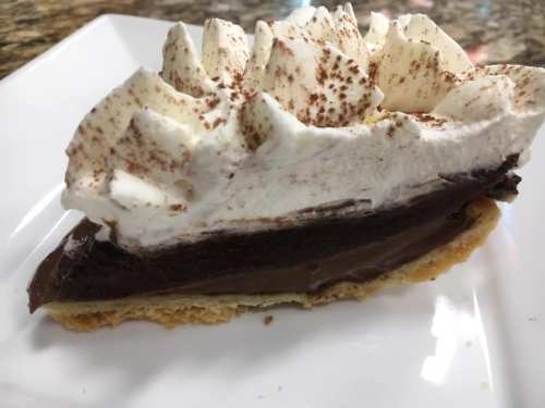 A slice of chocolate cream pie topped with whipped cream and cocoa powder on a white plate.
