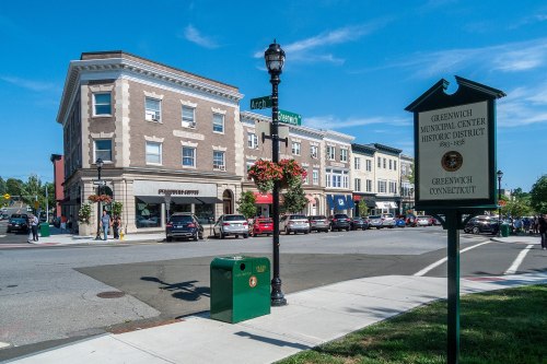 A sunny street view of Greenwich, Connecticut, featuring historic buildings, shops, and a municipal sign.