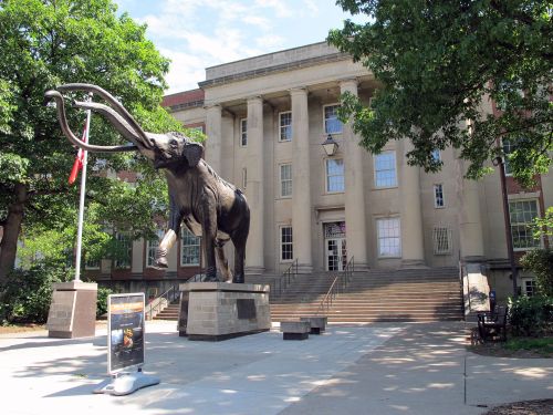 A large mammoth statue stands in front of a historic building with columns and steps, surrounded by trees.