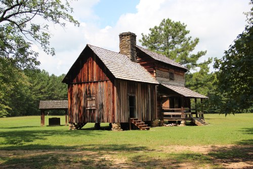 A rustic wooden house with a stone chimney, surrounded by green grass and trees under a partly cloudy sky.