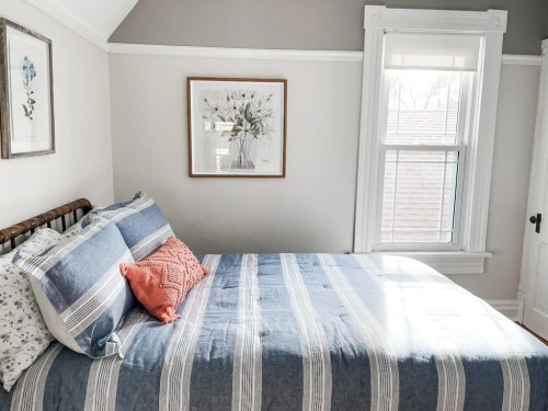 Cozy bedroom with a striped blue bedspread, decorative pillows, and framed artwork on the wall. Natural light from the window.