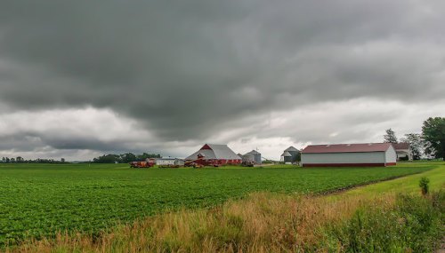 A rural landscape featuring a farm with red barns under a cloudy sky, surrounded by green fields.