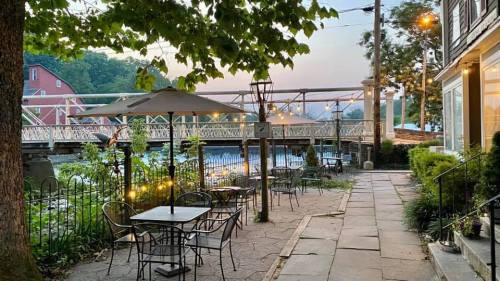 A serene outdoor dining area with tables, umbrellas, and a view of a bridge over a calm waterway at dusk.