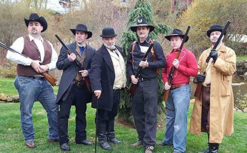 A group of six men dressed in Western attire, holding rifles, posing outdoors with trees in the background.