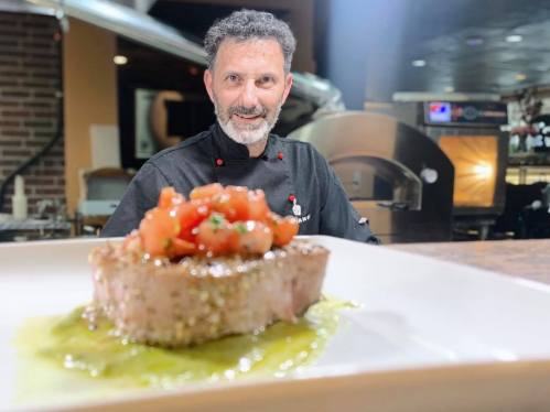 A chef smiles behind a beautifully plated dish of steak topped with fresh tomatoes and herbs in a restaurant setting.