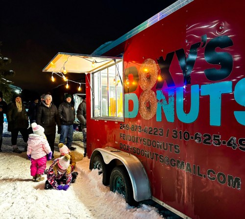 A red food truck named Doxy's Donuts with people gathered around in a snowy setting at night.