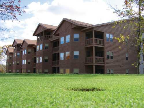 A row of brown wooden apartment buildings with balconies, set against a green lawn and blue sky.