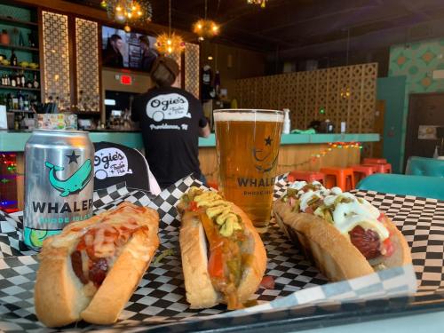 A tray of three gourmet hot dogs with toppings, a beer glass, and a can, set in a vibrant bar atmosphere.